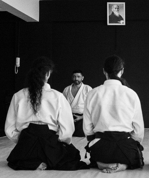 Dois praticantes de Aikido no dojo, de costas, segurando uma espada de madeira e um bastão